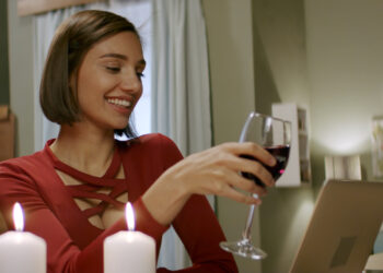 Beautiful woman in a red dress having a glass of wine chatting, online date with partner. Stay home, quarantine life. Shot with anamorphic lens